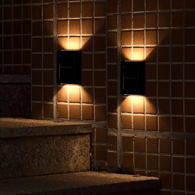SOLARIFY One solar-powered outdoor wall light mounted on a stone wall, projecting warm white light upward and downward. IP65 weatherproof. Suitable for garden fences, façades, covered terraces and entrance walls.