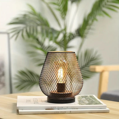 Ambientify Vira cordless metal mesh cage table lamp with a warm Edison-style bulb glowing through a diamond-pattern shade, placed on a wooden surface beside dried flowers and stacked books in a contemporary home interior.