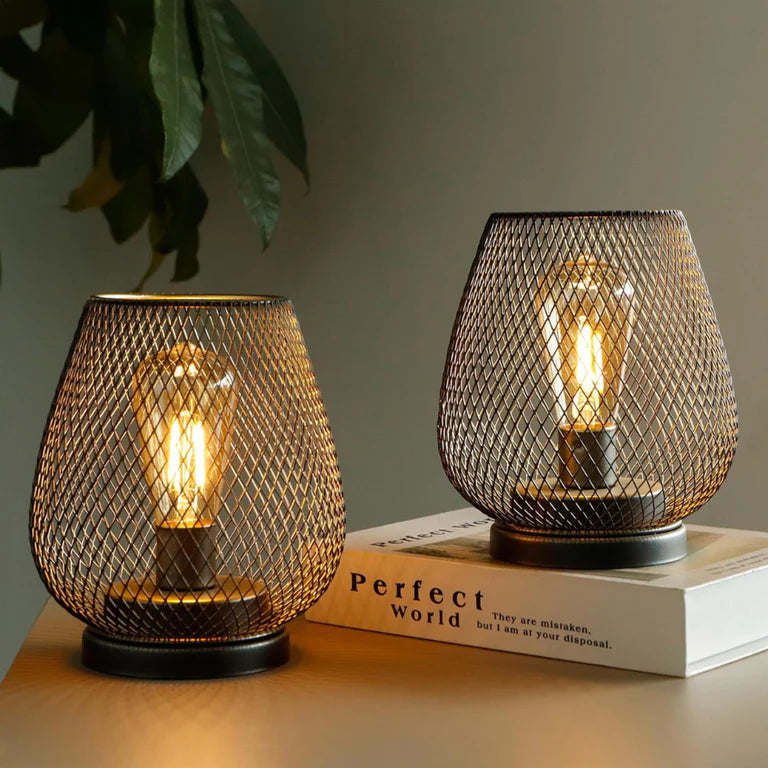 Ambientify Vira cordless metal mesh cage table lamp with a warm Edison-style bulb glowing through a diamond-pattern shade, placed on a wooden surface beside dried flowers and stacked books in a contemporary home interior.