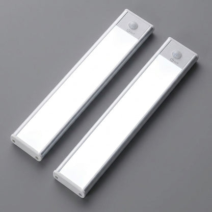AMBIENTIFY Sora slim rechargeable LED light bar mounted beneath a wooden shelf above a home desk, casting warm even light across the workspace — motion sensor under-cabinet lighting in brushed aluminium.