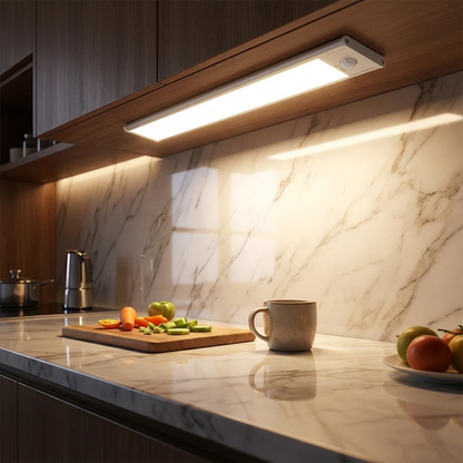 AMBIENTIFY Sora slim rechargeable LED light bar mounted beneath a wooden shelf above a home desk, casting warm even light across the workspace — motion sensor under-cabinet lighting in brushed aluminium.