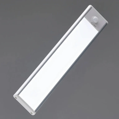 AMBIENTIFY Sora slim rechargeable LED light bar mounted beneath a wooden shelf above a home desk, casting warm even light across the workspace — motion sensor under-cabinet lighting in brushed aluminium.