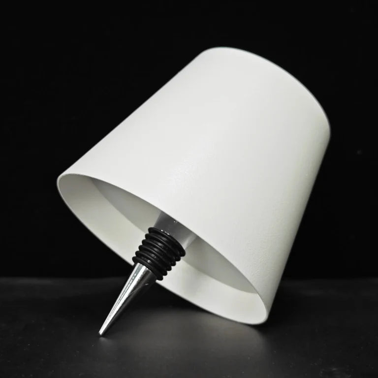 AMBIENTIFY Bottle wireless LED lamp fitted onto a wine bottle, placed on an outdoor garden table surrounded by natural greenery. Soft warm ambient glow, touch-dimmable in 3 levels, USB-C rechargeable table lamp for home décor.