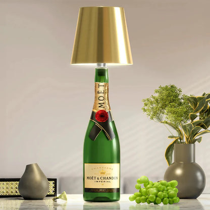AMBIENTIFY Bottle wireless LED lamp fitted onto a wine bottle, placed on an outdoor garden table surrounded by natural greenery. Soft warm ambient glow, touch-dimmable in 3 levels, USB-C rechargeable table lamp for home décor.