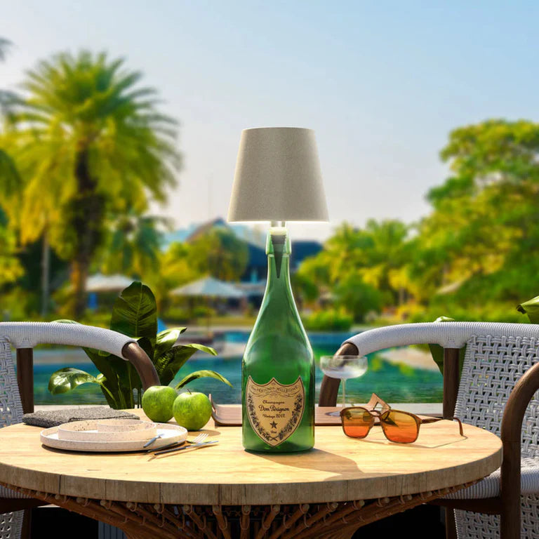 AMBIENTIFY Bottle wireless LED lamp fitted onto a wine bottle, placed on an outdoor garden table surrounded by natural greenery. Soft warm ambient glow, touch-dimmable in 3 levels, USB-C rechargeable table lamp for home décor.