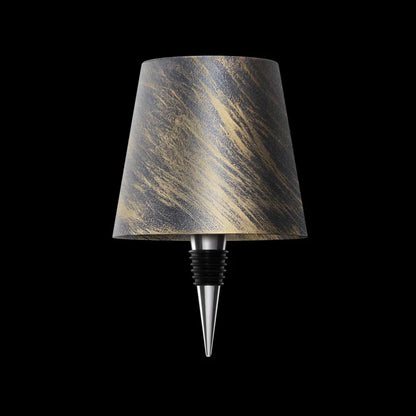 AMBIENTIFY Bottle wireless LED lamp fitted onto a wine bottle, placed on an outdoor garden table surrounded by natural greenery. Soft warm ambient glow, touch-dimmable in 3 levels, USB-C rechargeable table lamp for home décor.