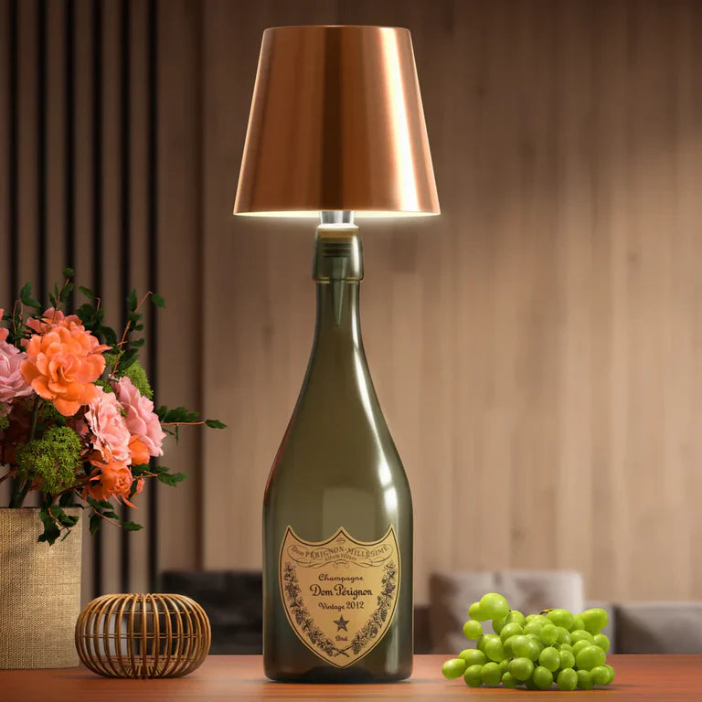 AMBIENTIFY Bottle wireless LED lamp fitted onto a wine bottle, placed on an outdoor garden table surrounded by natural greenery. Soft warm ambient glow, touch-dimmable in 3 levels, USB-C rechargeable table lamp for home décor.