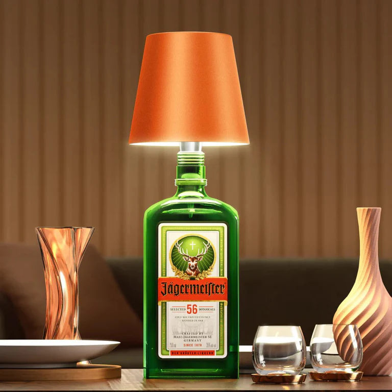 AMBIENTIFY Bottle wireless LED lamp fitted onto a wine bottle, placed on an outdoor garden table surrounded by natural greenery. Soft warm ambient glow, touch-dimmable in 3 levels, USB-C rechargeable table lamp for home décor.