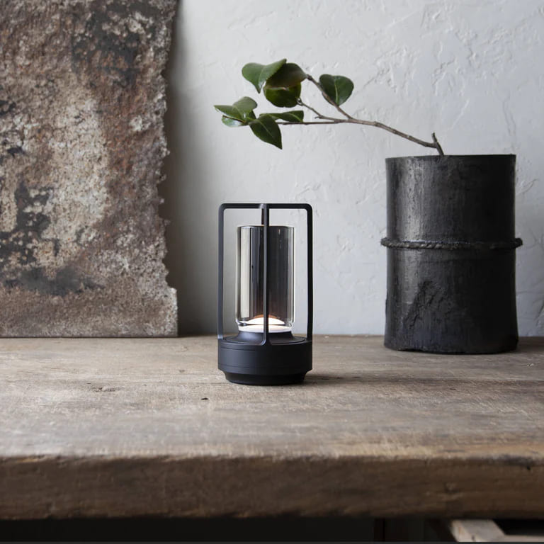 AMBIENTIFY Retro cordless retro table lantern lamp casting a warm amber glow, placed on a wooden tray. Slender aluminium frame with cylindrical acrylic glass shade. Atmospheric home décor lighting for bedroom, living room or outdoor use.