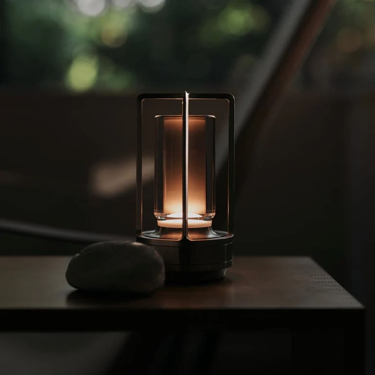 AMBIENTIFY Retro cordless retro table lantern lamp casting a warm amber glow, placed on a wooden tray. Slender aluminium frame with cylindrical acrylic glass shade. Atmospheric home décor lighting for bedroom, living room or outdoor use.