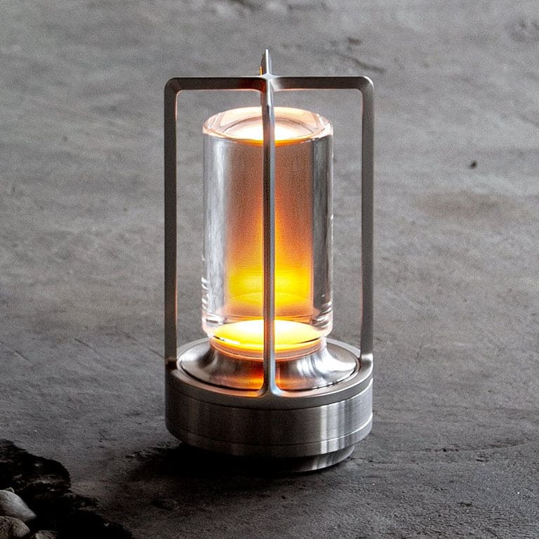 AMBIENTIFY Retro cordless retro table lantern lamp casting a warm amber glow, placed on a wooden tray. Slender aluminium frame with cylindrical acrylic glass shade. Atmospheric home décor lighting for bedroom, living room or outdoor use.