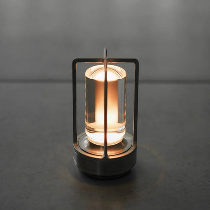 AMBIENTIFY Retro cordless retro table lantern lamp casting a warm amber glow, placed on a wooden tray. Slender aluminium frame with cylindrical acrylic glass shade. Atmospheric home décor lighting for bedroom, living room or outdoor use.