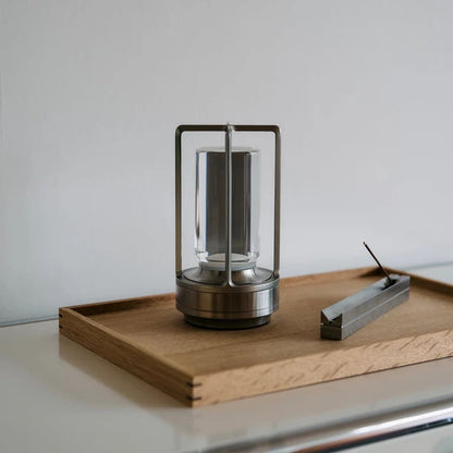 AMBIENTIFY Retro cordless retro table lantern lamp casting a warm amber glow, placed on a wooden tray. Slender aluminium frame with cylindrical acrylic glass shade. Atmospheric home décor lighting for bedroom, living room or outdoor use.