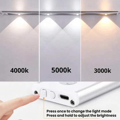 AMBIENTIFY Sora slim rechargeable LED light bar mounted beneath a wooden shelf above a home desk, casting warm even light across the workspace — motion sensor under-cabinet lighting in brushed aluminium.