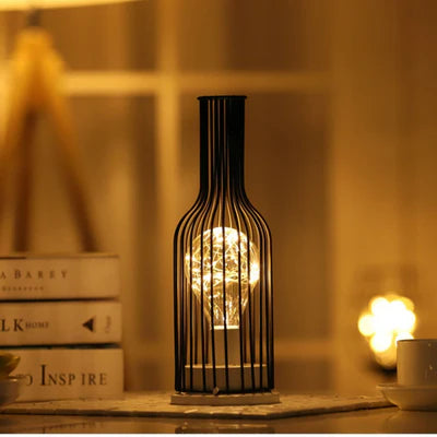 AMBIENTIFY Velora cordless wire-cage table lamp with warm amber LED glow placed on a wooden side table, casting golden light through a sculptural black steel frame — cable-free home accent lighting for any room.