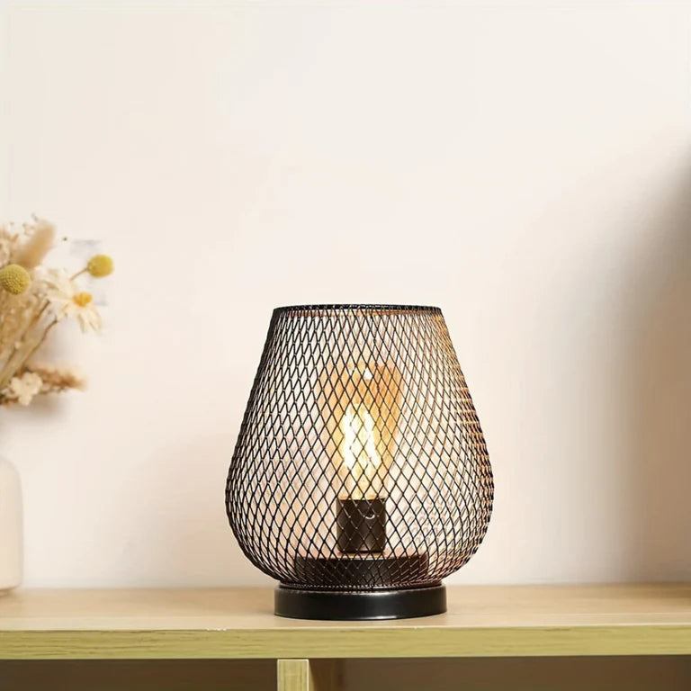 Ambientify Vira cordless metal mesh cage table lamp with a warm Edison-style bulb glowing through a diamond-pattern shade, placed on a wooden surface beside dried flowers and stacked books in a contemporary home interior.