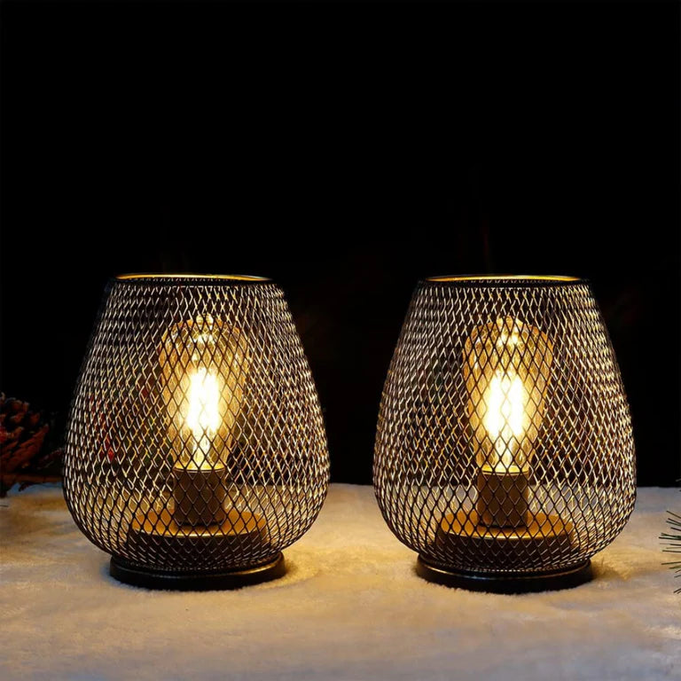 Ambientify Vira cordless metal mesh cage table lamp with a warm Edison-style bulb glowing through a diamond-pattern shade, placed on a wooden surface beside dried flowers and stacked books in a contemporary home interior.