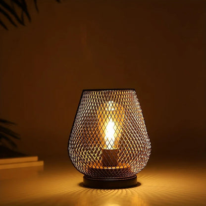 Ambientify Vira cordless metal mesh cage table lamp with a warm Edison-style bulb glowing through a diamond-pattern shade, placed on a wooden surface beside dried flowers and stacked books in a contemporary home interior.