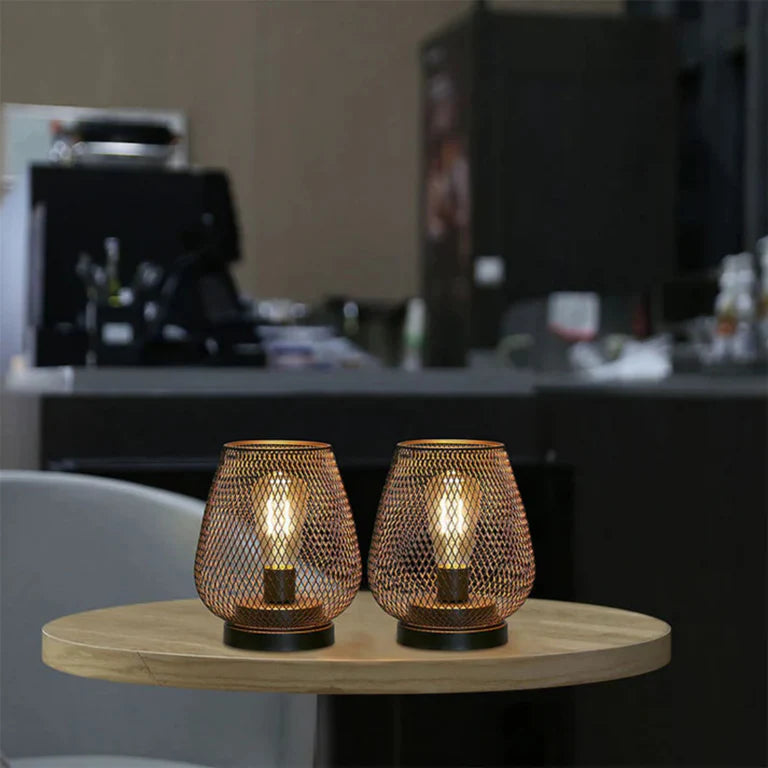 Ambientify Vira cordless metal mesh cage table lamp with a warm Edison-style bulb glowing through a diamond-pattern shade, placed on a wooden surface beside dried flowers and stacked books in a contemporary home interior.
