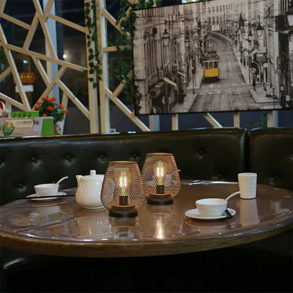 Ambientify Vira cordless metal mesh cage table lamp with a warm Edison-style bulb glowing through a diamond-pattern shade, placed on a wooden surface beside dried flowers and stacked books in a contemporary home interior.