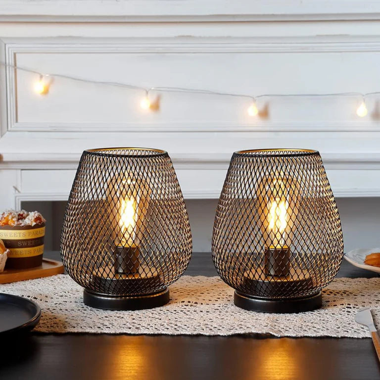 Ambientify Vira cordless metal mesh cage table lamp with a warm Edison-style bulb glowing through a diamond-pattern shade, placed on a wooden surface beside dried flowers and stacked books in a contemporary home interior.