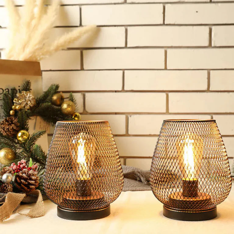 Ambientify Vira cordless metal mesh cage table lamp with a warm Edison-style bulb glowing through a diamond-pattern shade, placed on a wooden surface beside dried flowers and stacked books in a contemporary home interior.