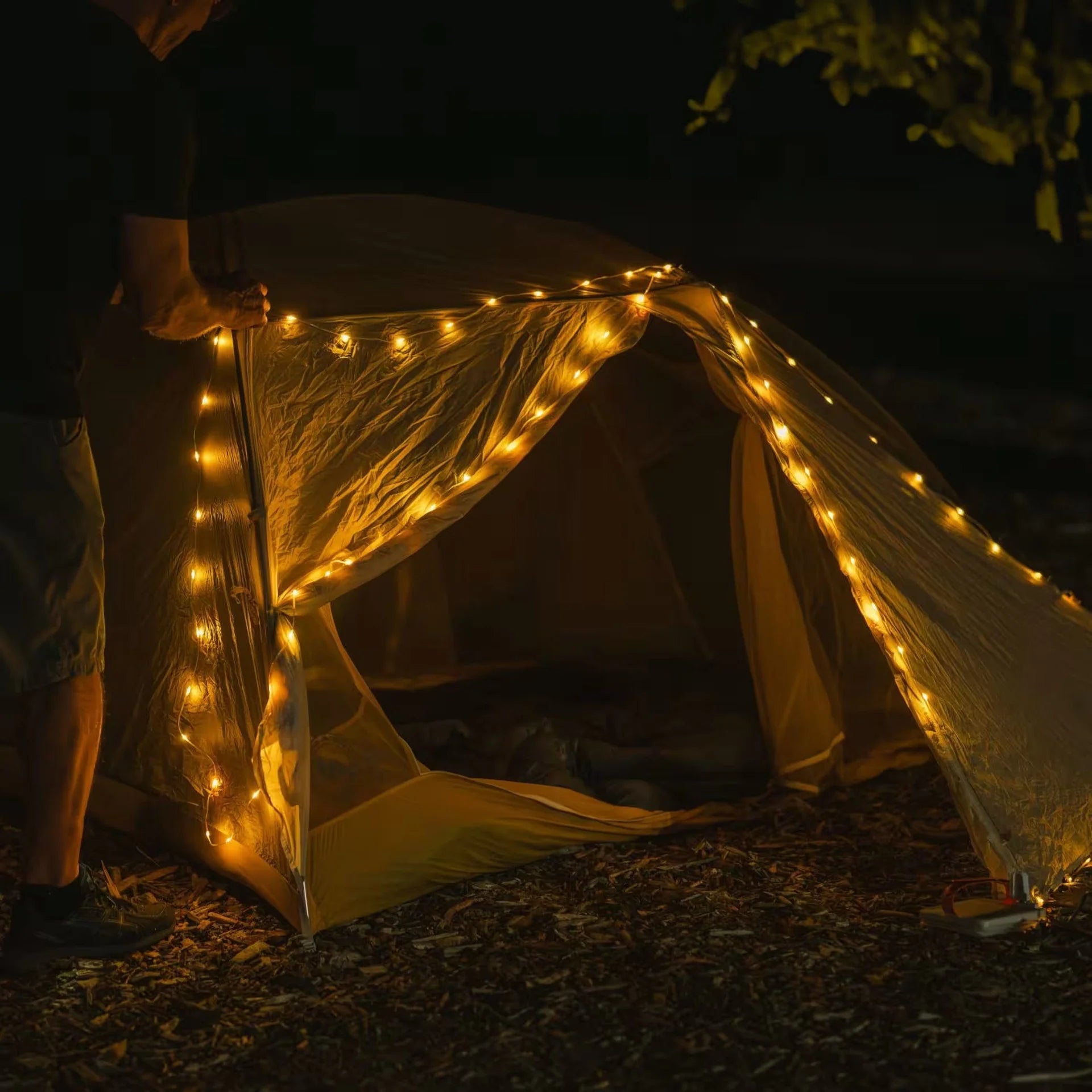 Campify String fairy light reel held in hand at night, showing warm amber LED string lights extended across dark outdoor ground, compact hub glowing with blue charge indicator, IP67 waterproof camping and home fairy lights.