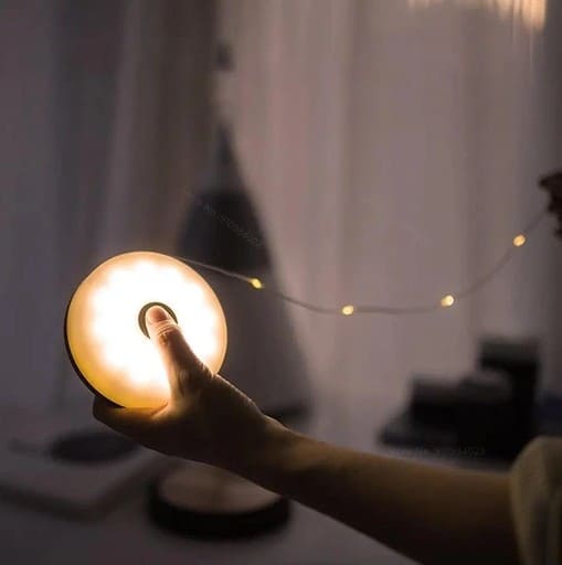 Campify String fairy light reel held in hand at night, showing warm amber LED string lights extended across dark outdoor ground, compact hub glowing with blue charge indicator, IP67 waterproof camping and home fairy lights.