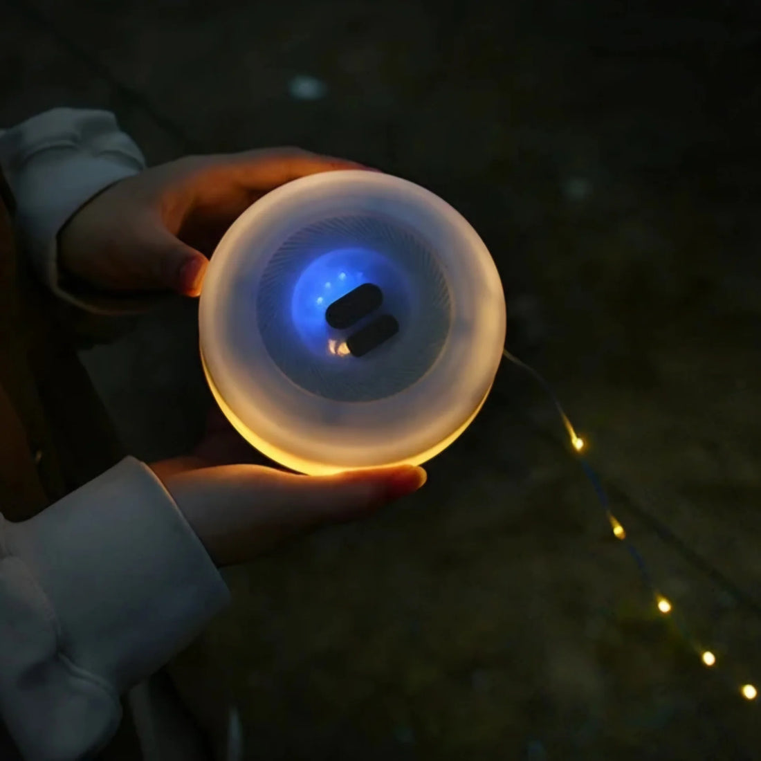 Campify String fairy light reel held in hand at night, showing warm amber LED string lights extended across dark outdoor ground, compact hub glowing with blue charge indicator, IP67 waterproof camping and home fairy lights.