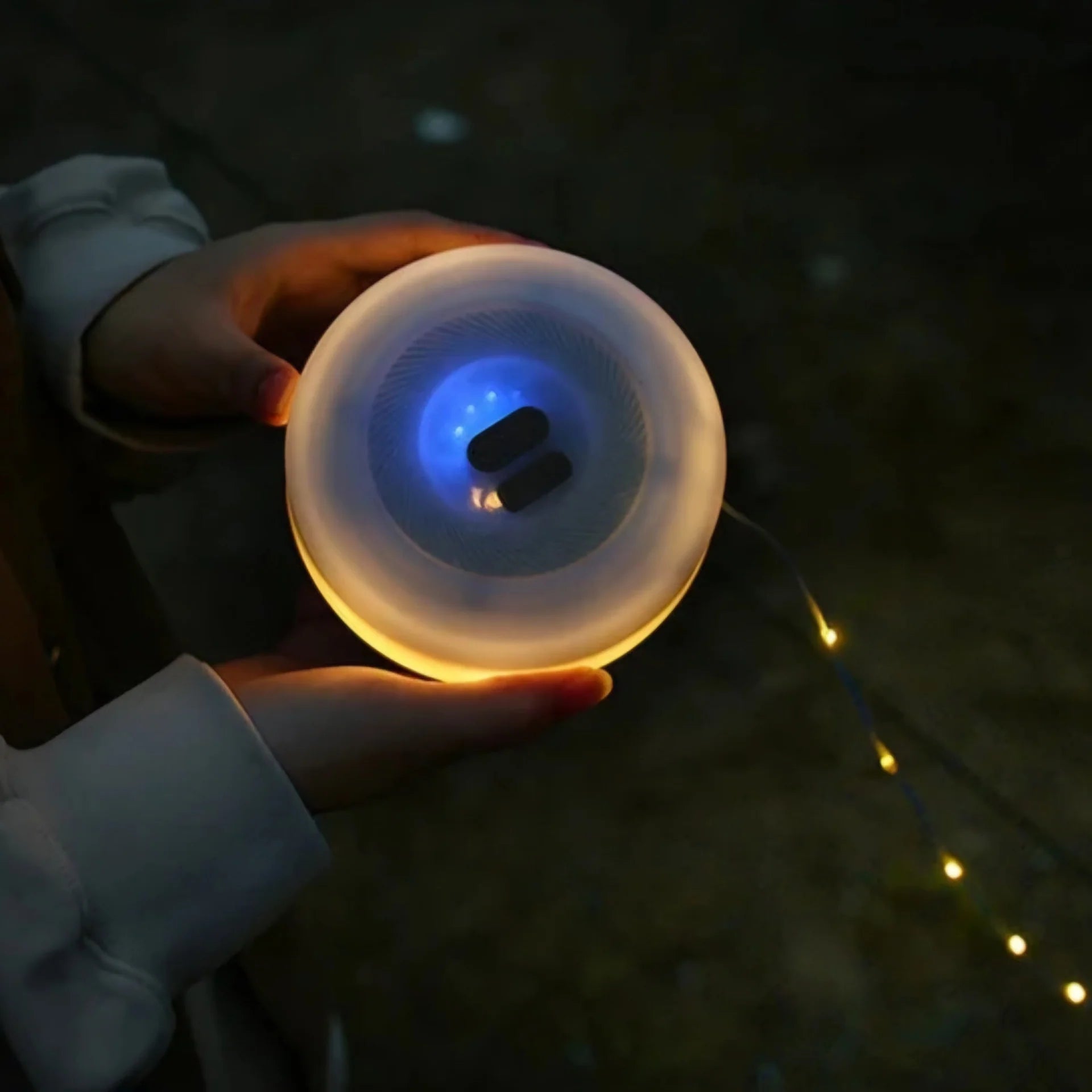 Campify String fairy light reel held in hand at night, showing warm amber LED string lights extended across dark outdoor ground, compact hub glowing with blue charge indicator, IP67 waterproof camping and home fairy lights.