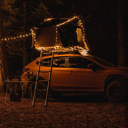 Campify String fairy light reel held in hand at night, showing warm amber LED string lights extended across dark outdoor ground, compact hub glowing with blue charge indicator, IP67 waterproof camping and home fairy lights.