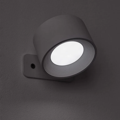 LUNIFY 360 wireless rechargeable LED spotlight with a circular concentric ring surround, mounted on a wall and casting warm amber light in a minimal interior — equally suited as a freestanding spotlight on a shelf or side table.