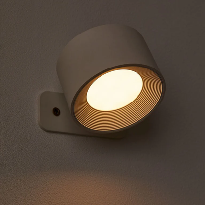 LUNIFY 360 wireless rechargeable LED spotlight with a circular concentric ring surround, mounted on a wall and casting warm amber light in a minimal interior — equally suited as a freestanding spotlight on a shelf or side table.