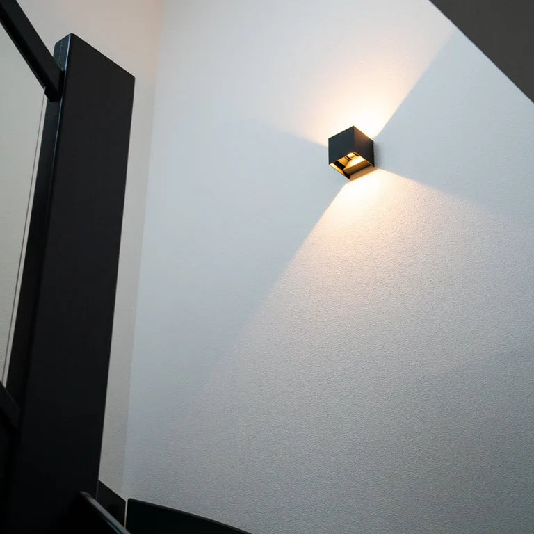 LUNIFY Boca compact aluminium wall light mounted on a slatted wood wall, casting a warm 2700K up-and-down glow. Wireless rechargeable LED wall lamp with motion sensor and magnetic mounting for wire-free indoor installation.