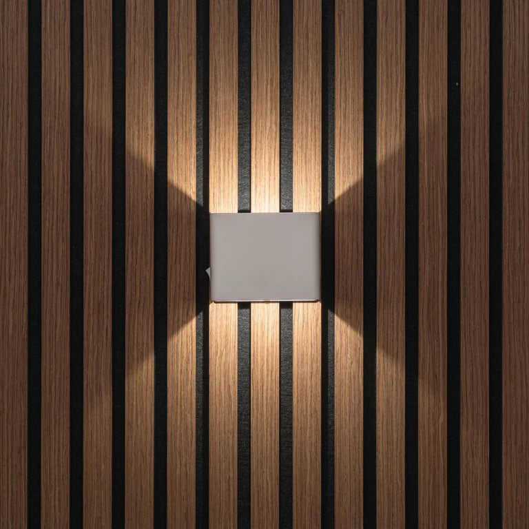 LUNIFY Boca compact aluminium wall light mounted on a slatted wood wall, casting a warm 2700K up-and-down glow. Wireless rechargeable LED wall lamp with motion sensor and magnetic mounting for wire-free indoor installation.