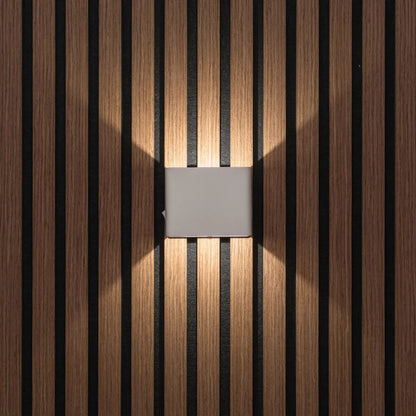 LUNIFY Boca compact aluminium wall light mounted on a slatted wood wall, casting a warm 2700K up-and-down glow. Wireless rechargeable LED wall lamp with motion sensor and magnetic mounting for wire-free indoor installation.