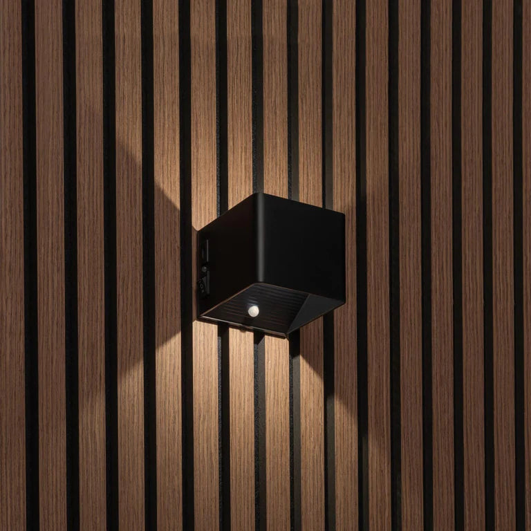 LUNIFY Boca compact aluminium wall light mounted on a slatted wood wall, casting a warm 2700K up-and-down glow. Wireless rechargeable LED wall lamp with motion sensor and magnetic mounting for wire-free indoor installation.