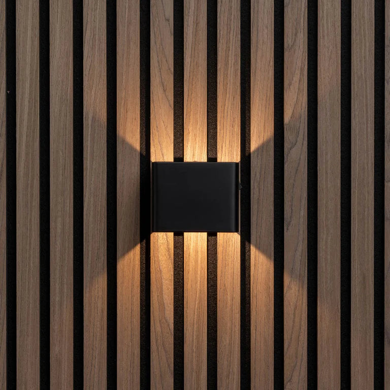 LUNIFY Boca compact aluminium wall light mounted on a slatted wood wall, casting a warm 2700K up-and-down glow. Wireless rechargeable LED wall lamp with motion sensor and magnetic mounting for wire-free indoor installation.