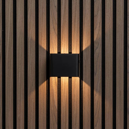 LUNIFY Boca compact aluminium wall light mounted on a slatted wood wall, casting a warm 2700K up-and-down glow. Wireless rechargeable LED wall lamp with motion sensor and magnetic mounting for wire-free indoor installation.