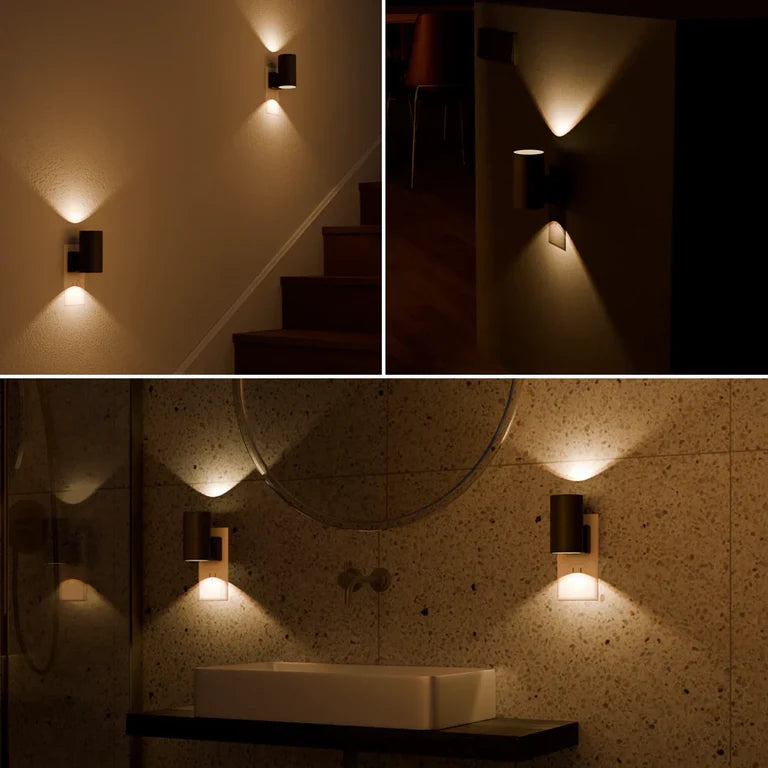 PLUGIFY One plug-in wall light in matte finish mounted in a bedroom wall socket, casting warm ambient light onto a wooden bedside table with a remote control and ceramic mug beside it.