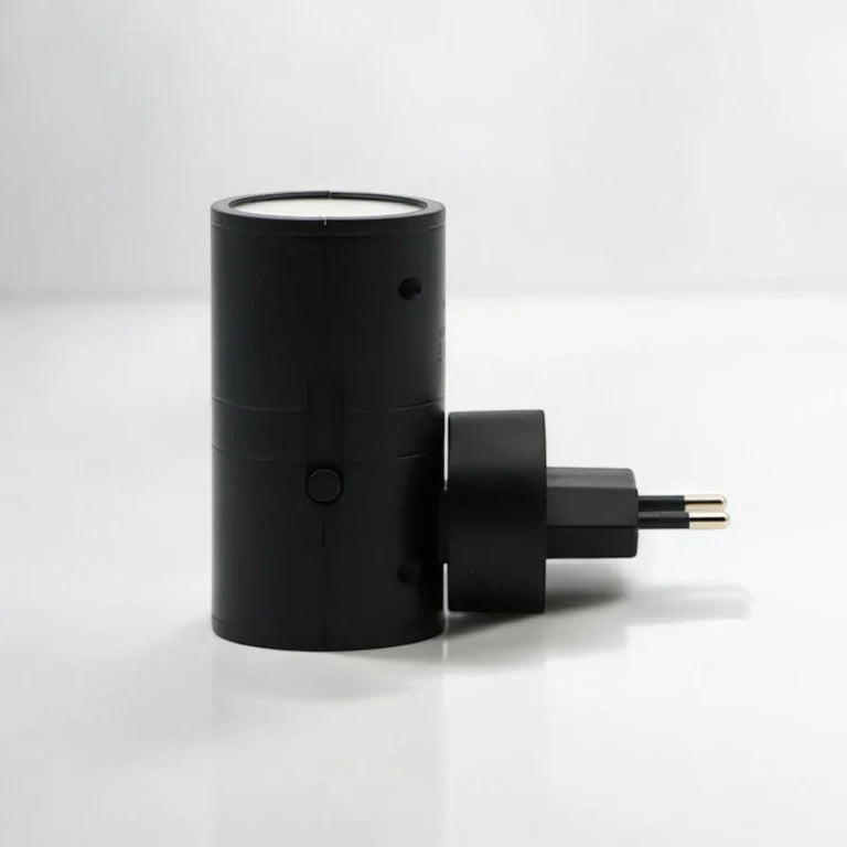 PLUGIFY One plug-in wall light in matte finish mounted in a bedroom wall socket, casting warm ambient light onto a wooden bedside table with a remote control and ceramic mug beside it.