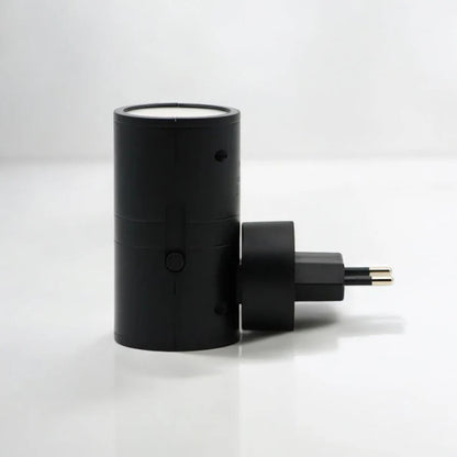 PLUGIFY One plug-in wall light in matte finish mounted in a bedroom wall socket, casting warm ambient light onto a wooden bedside table with a remote control and ceramic mug beside it.