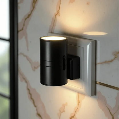 PLUGIFY One plug-in wall light in matte finish mounted in a bedroom wall socket, casting warm ambient light onto a wooden bedside table with a remote control and ceramic mug beside it.