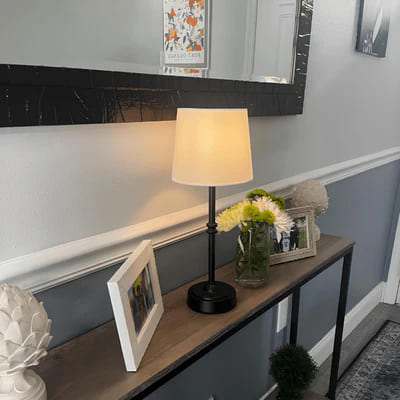 AMBIENTIFY Classic cordless table lamp with fabric shade and iron stem, casting warm ambient light on a bedside table. Three brightness settings and up to 40 hours battery life. Suitable for bedroom, living room and home office.