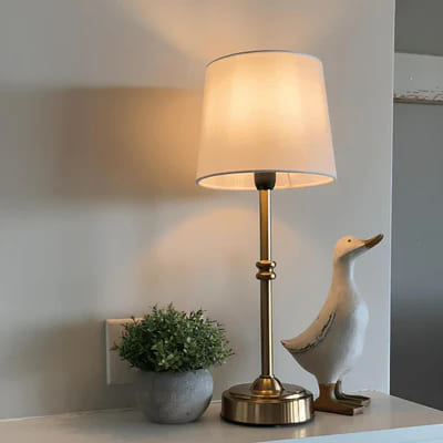 AMBIENTIFY Classic cordless table lamp with fabric shade and iron stem, casting warm ambient light on a bedside table. Three brightness settings and up to 40 hours battery life. Suitable for bedroom, living room and home office.