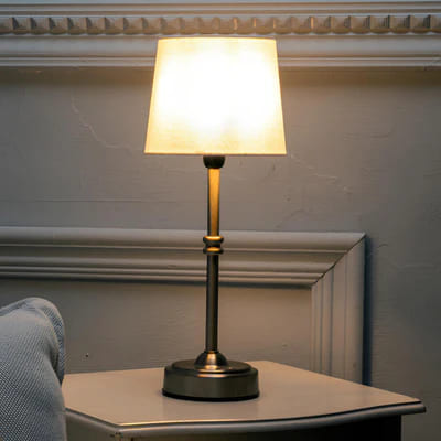 AMBIENTIFY Classic cordless table lamp with fabric shade and iron stem, casting warm ambient light on a bedside table. Three brightness settings and up to 40 hours battery life. Suitable for bedroom, living room and home office.