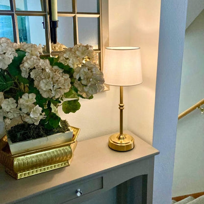 AMBIENTIFY Classic cordless table lamp with fabric shade and iron stem, casting warm ambient light on a bedside table. Three brightness settings and up to 40 hours battery life. Suitable for bedroom, living room and home office.