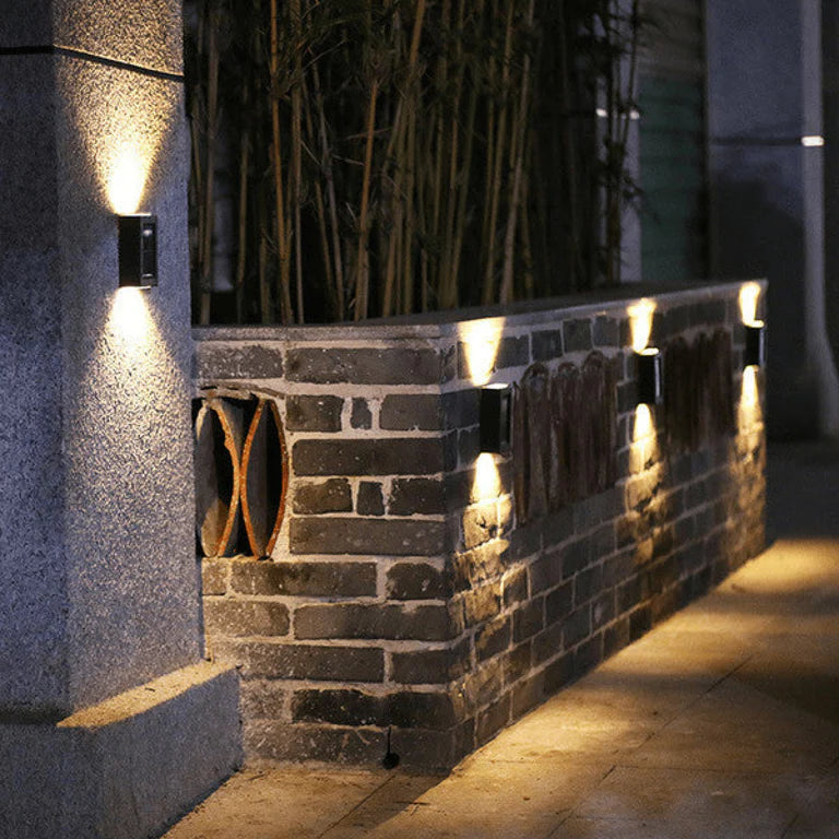 SOLARIFY One solar-powered outdoor wall light mounted on a stone wall, projecting warm white light upward and downward. IP65 weatherproof. Suitable for garden fences, façades, covered terraces and entrance walls.